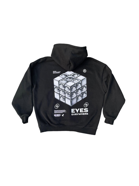 The "[Pre-order] Eyes Everywhere Hoodie" from Snaked is a trendy black hoodie made of premium materials, showcasing a unique Rubik's cube design adorned with eyes on each square on the back. The phrase "Eyes Everywhere" is printed beneath the design, surrounded by an assortment of small symbols and text elements.